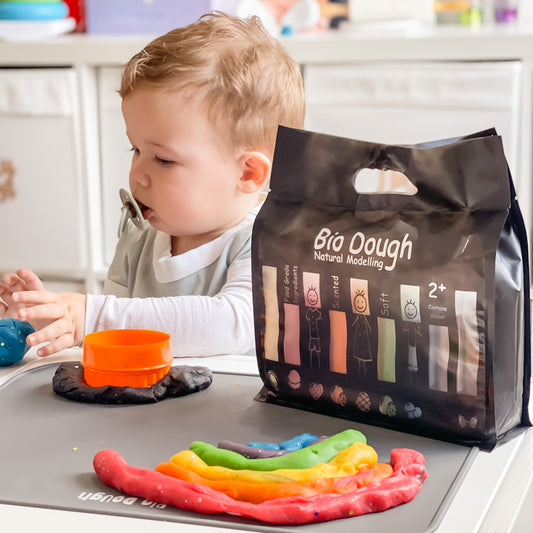 Bio Dough Natural Modelling clay kit in a black resealable bag, surrounded by multiple colorful dough balls in white, pink, red, orange, yellow, green, blue, and purple on a peach background.