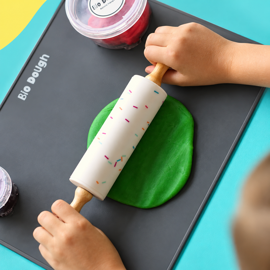 A dark gray silicone baking mat with rounded corners, featuring the white text 'Bio Dough' near the top and a small star-shaped cutout in the upper right corner, displayed on a colorful background of yellow, light blue, and mint green shapes.