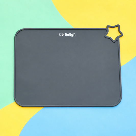 A dark gray silicone baking mat with rounded corners, featuring the white text 'Bio Dough' near the top and a small star-shaped cutout in the upper right corner, displayed on a colorful background of yellow, light blue, and mint green shapes.