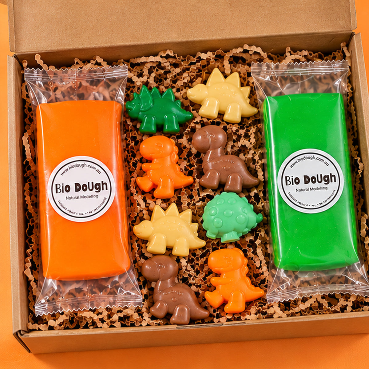 a close up of a dinosaur playdough kit with orange and green playdough pouches and dinosaur figurines in yellow, brown, green and aqua.